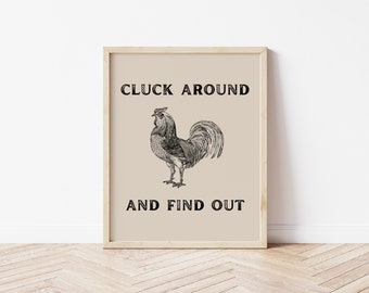 Puede incluir: Impresión enmarcada con una ilustración en blanco y negro de un gallo. El texto "CLUCK AROUND AND FIND OUT" se muestra en letras mayúsculas negras y en negrita. El fondo es beige y el marco es de color madera clara.