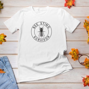May include: White t-shirt with a black circle graphic that says "Bee Sting Survivor" with a bee illustration inside the circle.