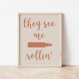以下が含まれることがあります： 茶色の筆記体で「they see me rollin'」と書かれた額装されたプリント。 「me」と「rollin'」の間に、茶色のシルエットの麺棒が中央に配置されています。
