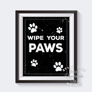 May include: Black and white sign with the text "Wipe Your Paws" in a distressed font. The sign is surrounded by four white paw prints.