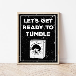 May include: Black and white printable wall art with a distressed look. The text reads "Let's get ready to tumble" with a graphic of a washing machine.