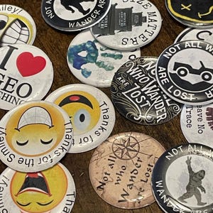May include: A collection of round pinback buttons with various designs and text. Some buttons feature smiley faces, a compass, a jeep, and the phrase "Not all who wander are lost." Other buttons have the text "Size Matters" and "I love geo caching".
