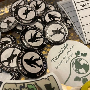 May include: A selection of black and white badges featuring the text "NOT ALL WHO WANDER ARE LOST" around a silhouette of a person. Also, a sticker with the text "Tamis2go" and a notebook with the text "NAME/DATE".