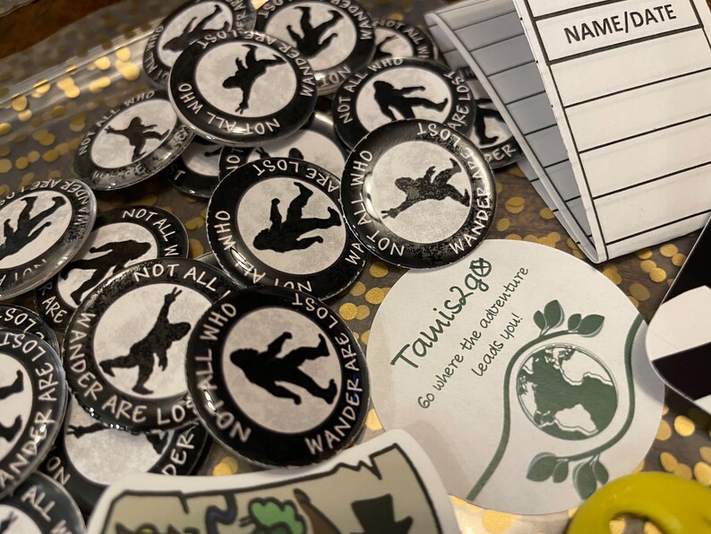 May include: A selection of black and white badges featuring the text "NOT ALL WHO WANDER ARE LOST" around a silhouette of a person. Also, a sticker with the text "Tamis2go" and a notebook with the text "NAME/DATE".