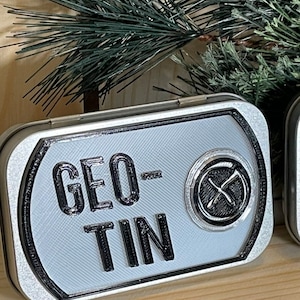 Può includere: Una scatola di metallo argentata con un design a bussola bianco e nero e il testo "GEO-TIN" stampato sul davanti.