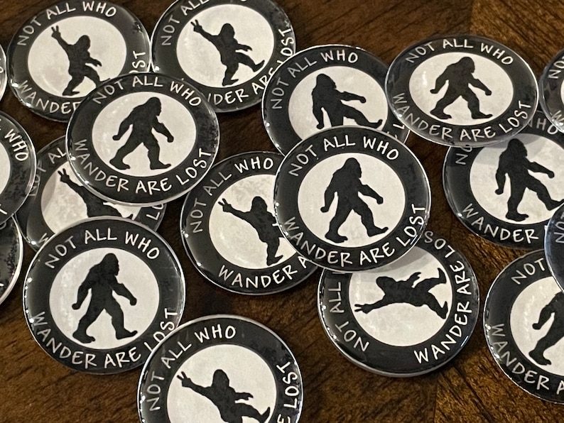 May include: A selection of black and white Bigfoot-themed badges. Each badge displays a black Bigfoot silhouette and the text "NOT ALL WHO WANDER ARE LOST" in a circular arrangement. The badges are arranged on a wooden surface.