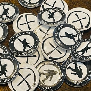 May include: A collection of black and white badges with the text "NOT ALL WHO WANDER ARE LOST". The badges feature a silhouette of a Bigfoot-like creature. Some badges have a cross design.