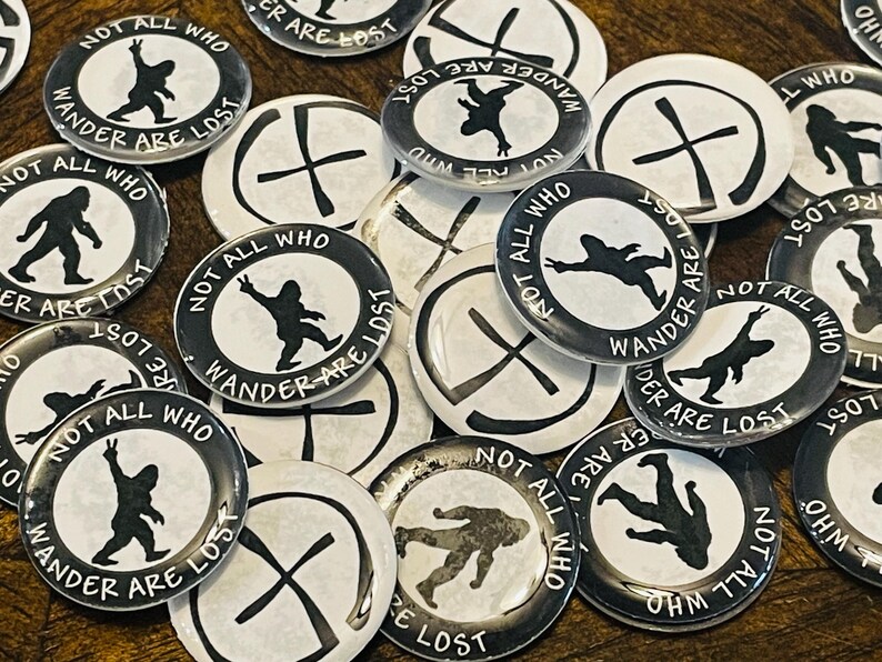 May include: A collection of black and white badges with the text "NOT ALL WHO WANDER ARE LOST". The badges feature a silhouette of a Bigfoot-like creature. Some badges have a cross design.