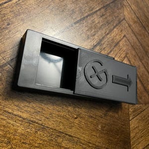 May include: A black, rectangular, sliding-top container. The container has a circular symbol with a cross and an arrow pointing to the right. The container is open, revealing a dark interior.