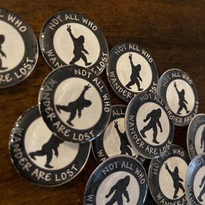 May include: A selection of circular black and white badges featuring a Bigfoot design and the words "NOT ALL WHO WANDER ARE LOST". The Bigfoot illustration is shown in different poses, some with a peace sign. The badges are arranged on a wooden surface.