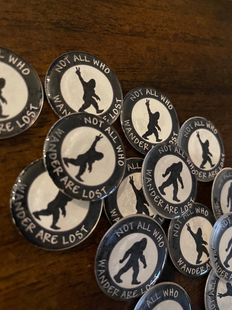 May include: A selection of circular black and white badges featuring a Bigfoot design and the words "NOT ALL WHO WANDER ARE LOST". The Bigfoot illustration is shown in different poses, some with a peace sign. The badges are arranged on a wooden surface.