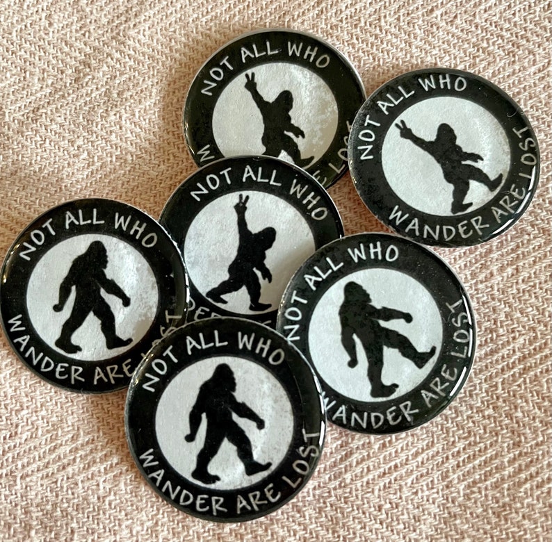 May include: A group of black and white Bigfoot-themed badges. Each badge displays a Bigfoot silhouette within a white circle, encircled by the text "NOT ALL WHO WANDER ARE LOST". The badges are arranged on a pink fabric background.
