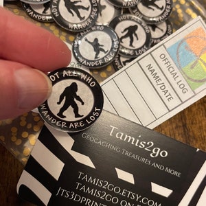 May include: A selection of black and white Bigfoot-themed badges, each with the phrase "Not All Who Wander Are Lost." The badges feature a Bigfoot silhouette and are likely for geocaching.