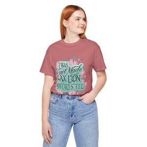 May include: A dusty rose t-shirt with the text "I was not made of ax iron and sword steel" in a teal and floral design. The shirt is made of a soft material and is suitable for casual wear.