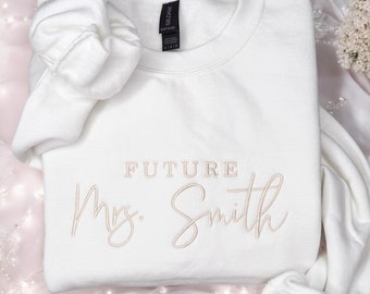 May include: A white crewneck sweatshirt with the words "FUTURE Mrs. Smith" embroidered in a light brown thread. The sweatshirt is folded, with one sleeve rolled up. The background is a soft, pinkish fabric.