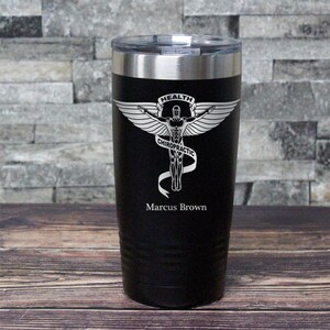 May include: Black stainless steel tumbler with a silver engraved design of a winged figure with the text "HEALTH CHIROPRACTIC" and the name "Marcus Brown".