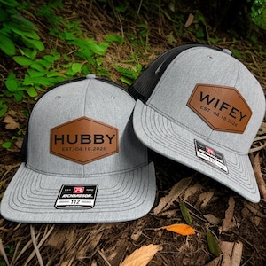 May include: Two gray trucker hats with black mesh backs. Each hat features a brown leather patch with the words "HUBBY" and "WIFEY" respectively, along with an established date. The hats have a curved brim and adjustable strap.