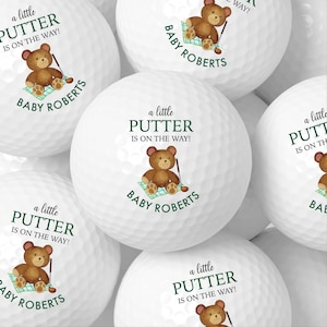 May include: White golf balls featuring a teddy bear graphic, a golf club, and the text "a little PUTTER IS ON THE WAY!" and "BABY ROBERTS". The golf balls are themed for a baby shower or gender reveal.