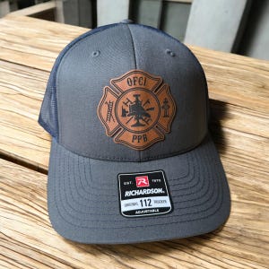 May include: A charcoal gray trucker hat with a navy blue mesh back and a brown leather patch on the front. The patch features a fire department emblem with the text OFCI and PPB. A black sticker on the brim reads Richardson Original 112 Trucker.