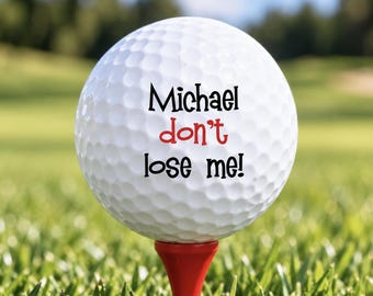 May include: A white golf ball with a dimpled surface sits on a red tee. The ball features black text that reads Michael and lose me! with the word don't printed in red text between them. The design is centered on the front of the golf ball.
