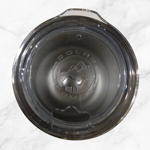 May include: Close-up of a stainless steel travel mug with a clear lid. The mug has a black logo that says "Polar Camel" with a camel silhouette and mountain range graphic.