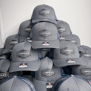 May include: A stack of grey trucker hats with navy blue mesh backs. Each hat features a black hexagonal patch on the front with the white text COSPOLICH and Made in the USA. A small rectangular Richardson 112 sticker is attached to the brim of each hat.