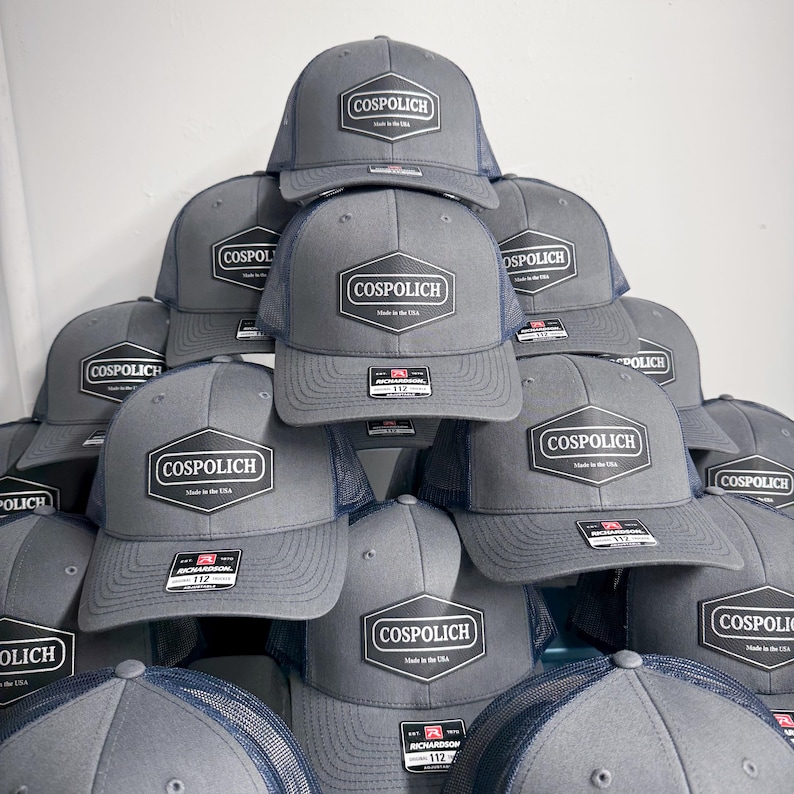 May include: A stack of grey trucker hats with navy blue mesh backs. Each hat features a black hexagonal patch on the front with the white text COSPOLICH and Made in the USA. A small rectangular Richardson 112 sticker is attached to the brim of each hat.