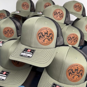 May include: A collection of olive green and black mesh trucker hats featuring a circular tan leather patch on the front. The patch displays an octopus graphic with the text JAYS BACHELOR PARTY 2023 FORT LAUDERDALE. Each hat has a Richardson 112 label.