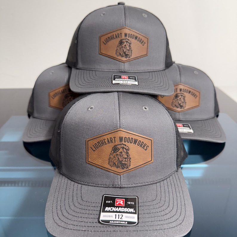 May include: A stack of gray trucker hats with black mesh backs. Each hat features a brown hexagonal leather patch on the front with a lion illustration and the text LIONHEART WOODWORKS. A black and white Richardson 112 sticker is on the brim.