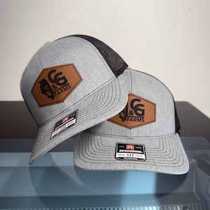 May include: Two grey and black trucker hats featuring a brown leather-like hexagonal patch on the front. The patch displays the text C&G FARMS with a state silhouette and corn stalk graphic. Each hat has a Richardson 112 adjustable sticker on the brim.