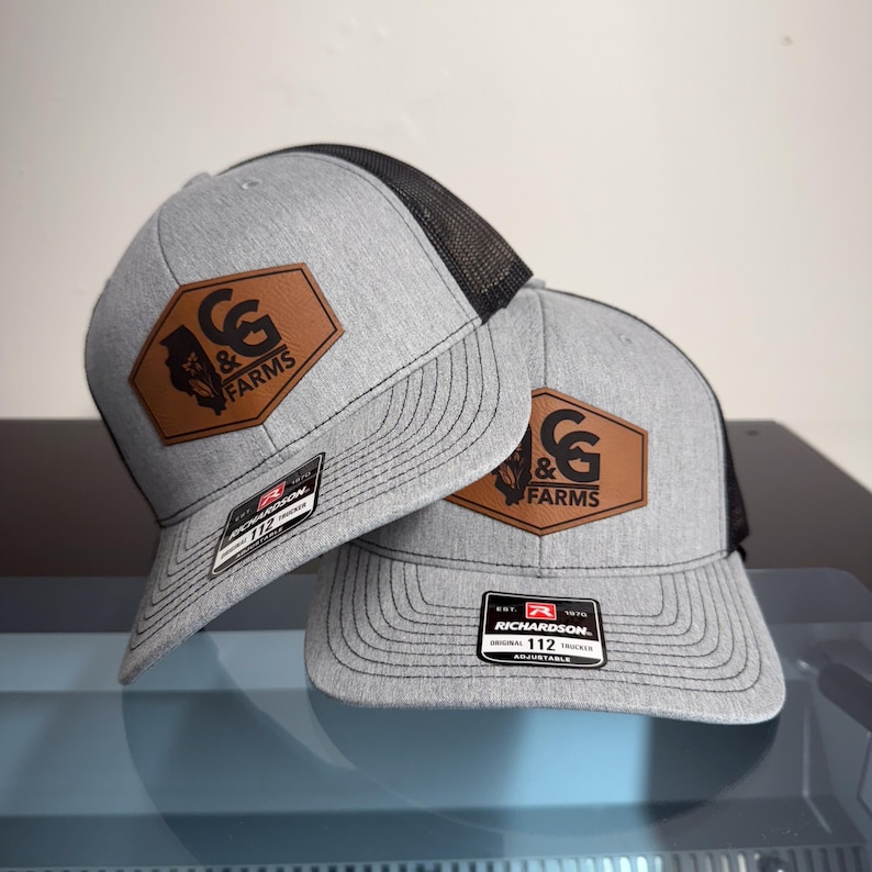 May include: Two grey and black trucker hats featuring a brown leather-like hexagonal patch on the front. The patch displays the text C&G FARMS with a state silhouette and corn stalk graphic. Each hat has a Richardson 112 adjustable sticker on the brim.