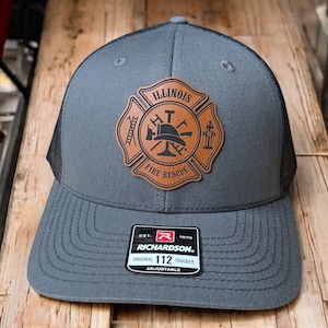 May include: A charcoal gray trucker hat featuring a black mesh back and a tan leather patch on the front. The patch is shaped like a Maltese cross with the text ILLINOIS FIRE RESCUE and icons of a ladder, hydrant, and helmet. A black sticker is on the brim.