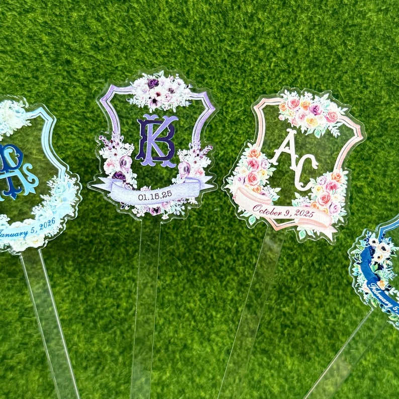 Op de afbeelding: Transparante acryl bruiloftsdecoraties met bloemmotieven en initialen. Elk stuk heeft een unieke initiaal, datum en bloemstuk in tinten paars, roze en blauw. De items staan op een groene achtergrond.