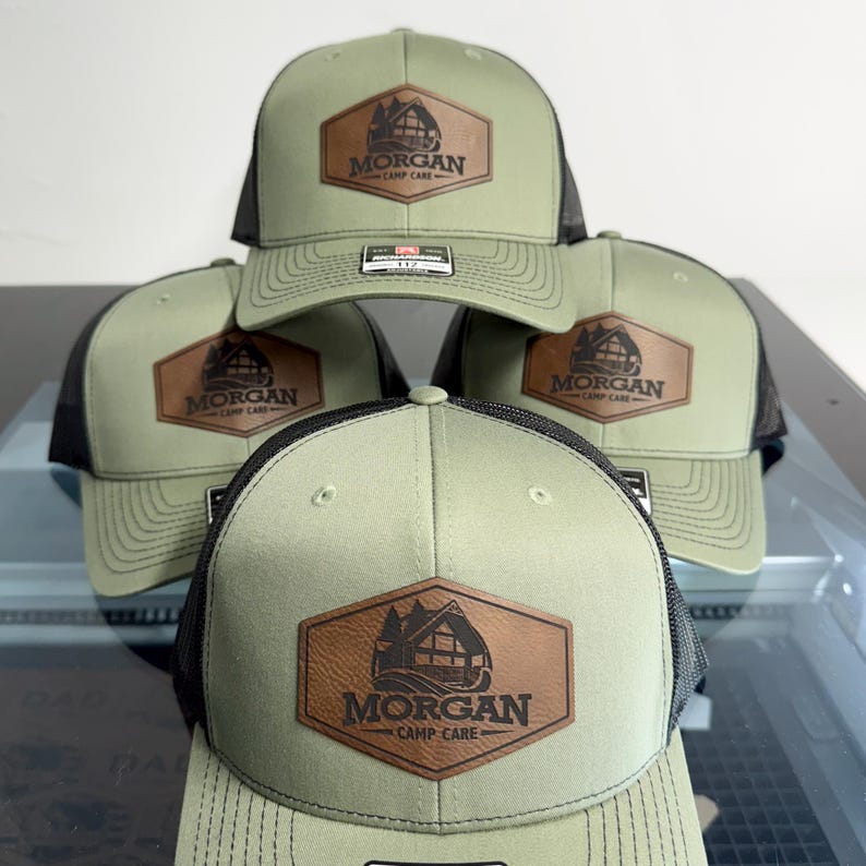 May include: A group of four olive green and black trucker hats featuring a brown hexagonal leather patch on the front. The patch displays a graphic of a cabin and the text Morgan Camp Care. Each hat has a structured crown and mesh back.