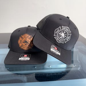 May include: Two black trucker hats with mesh backs and adjustable snapback closures. Each hat features a Richardson 112 label on the brim and a circular fire department emblem on the front. One emblem reads City of Northville, the other Detroit Fire Dept.
