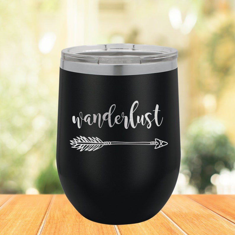 May include: Black stainless steel wine tumbler with a silver lid. The tumbler has the word "wanderlust" and an arrow engraved on the side.