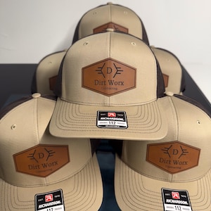 May include: A stack of tan and brown trucker hats featuring a brown hexagonal leather patch on the front. The patch displays the text Dirt Worx Contracting with a stylized letter D. Each hat includes a Richardson 112 adjustable sticker.