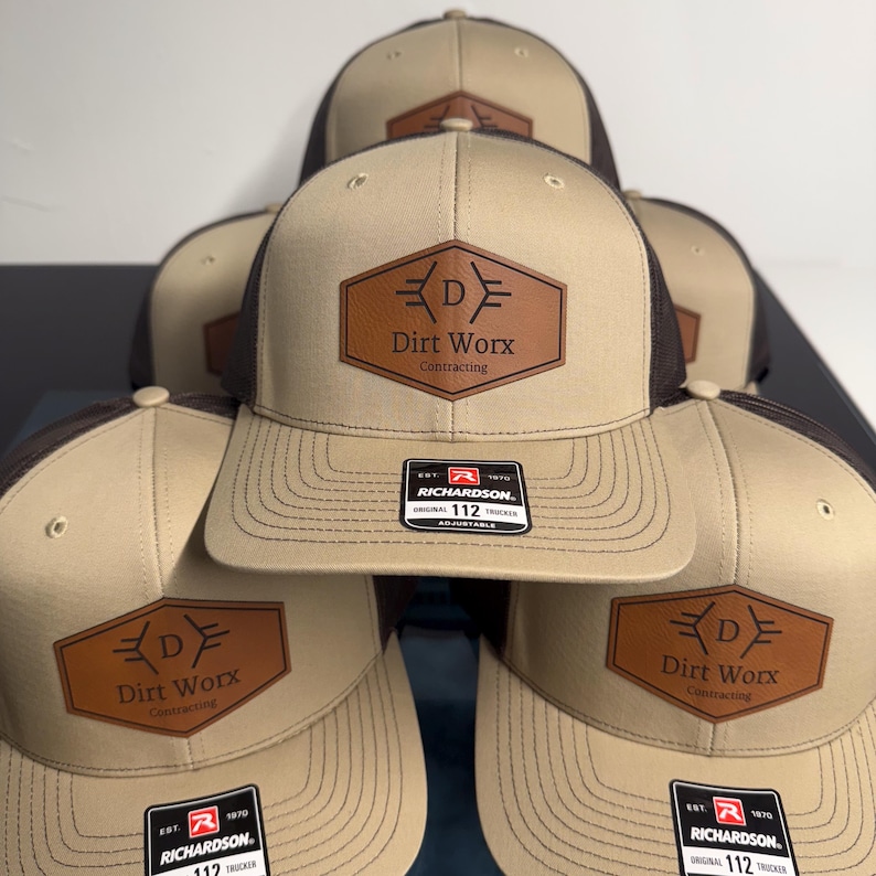 May include: A stack of tan and brown trucker hats featuring a brown hexagonal leather patch on the front. The patch displays the text Dirt Worx Contracting with a stylized letter D. Each hat includes a Richardson 112 adjustable sticker.