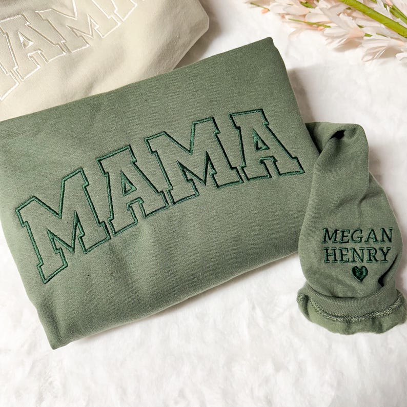 May include: A green sweatshirt with the word "MAMA" embroidered in a varsity style font. A smaller green garment with the text "MEGAN HENRY" and a heart is also shown.