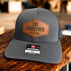 May include: A gray trucker hat with a brown leather patch that reads "THE GODFATHER of Bobby." The hat has a mesh back and an adjustable strap. A small white tag with black text is on the front.