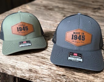 May include: Two trucker hats with a brown leather patch that says "Made in 1945" and "Vintage 1945 All Original Parts". The hats are green and gray with black mesh.