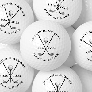 May include: White golf balls with black text and graphics. Each ball has the text "IN LOVING MEMORY", the years "1949 2024", and the name "MARK A. BANKS". The design includes crossed golf clubs and a golf ball.