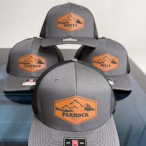 May include: Four gray trucker hats with black mesh backs. Each hat features a hexagonal tan patch with a mountain and forest graphic. The patches are personalized with the names DEVITT, CASTILLO, BELLA, and PARROCK printed in black text.