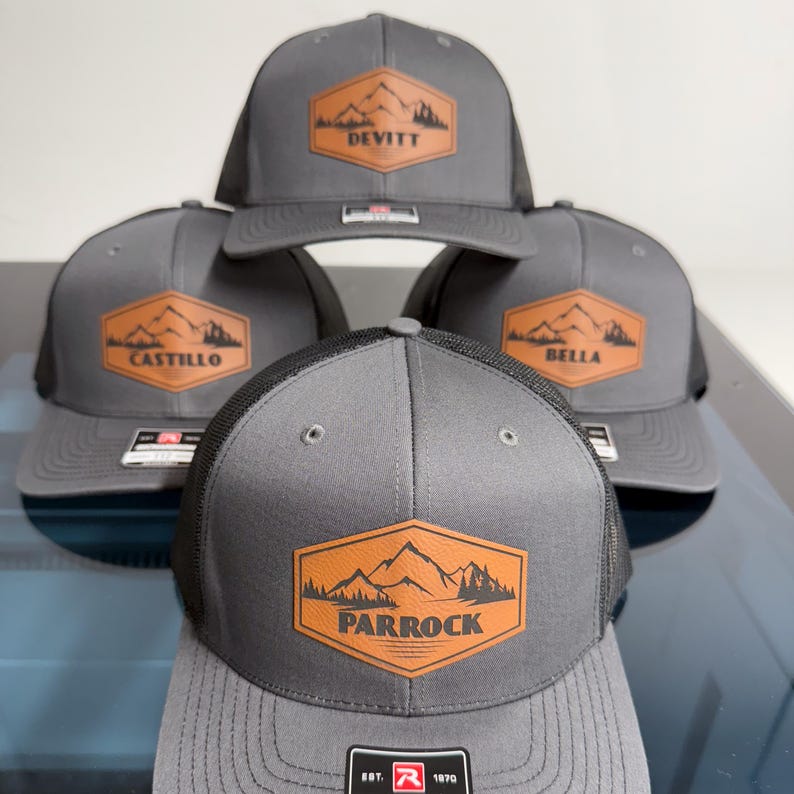 May include: Four gray trucker hats with black mesh backs. Each hat features a hexagonal tan patch with a mountain and forest graphic. The patches are personalized with the names DEVITT, CASTILLO, BELLA, and PARROCK printed in black text.