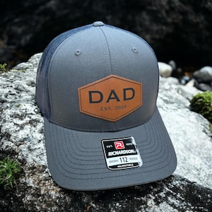 May include: A gray and black trucker hat with a brown leather patch that says "DAD EST. 2024". The hat is adjustable and has a Richardson 112 label.