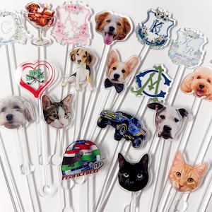Op de afbeelding: Een verzameling heldere acryl cocktailroerstaafjes met verschillende huisdierportretten, waaronder honden en katten, en andere ontwerpen. Sommige roerstaafjes hebben decoratieve elementen zoals bloemenwapens en een raceautohelm.