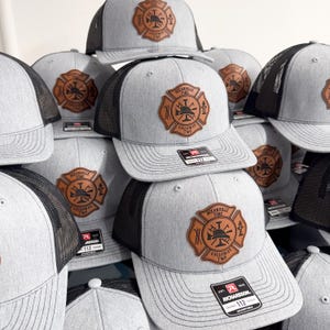 May include: A collection of grey and black trucker hats featuring a brown leather patch on the front. The patch is shaped like a Maltese cross and displays the text Oceanville Fire Galloway Twp along with icons of a fire helmet and ladder.