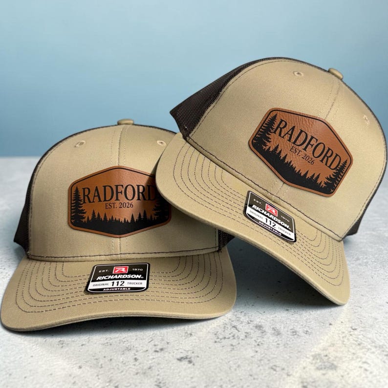 May include: Two tan and brown trucker hats featuring a brown leather-like patch on the front. The patch displays a black silhouette of pine trees and the text RADFORD EST. 2026. Each hat has a Richardson 112 adjustable sticker on the brim.