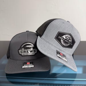 May include: Two trucker hats with mesh backs. The left hat is dark gray with a black patch featuring a car graphic and the text CREWS AUTOMOTIVE. The right hat is heather gray with a black patch featuring a house graphic and the text CAGE'S CARPENTRY.