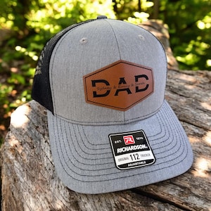 Peut inclure: Un chapeau de camionneur gris et noir avec un patch en cuir marron qui dit "DAD" avec les noms "Sydney, Emily & Sarah" en dessous. Le chapeau a un dos en maille noire et un logo Richardson sur le côté.